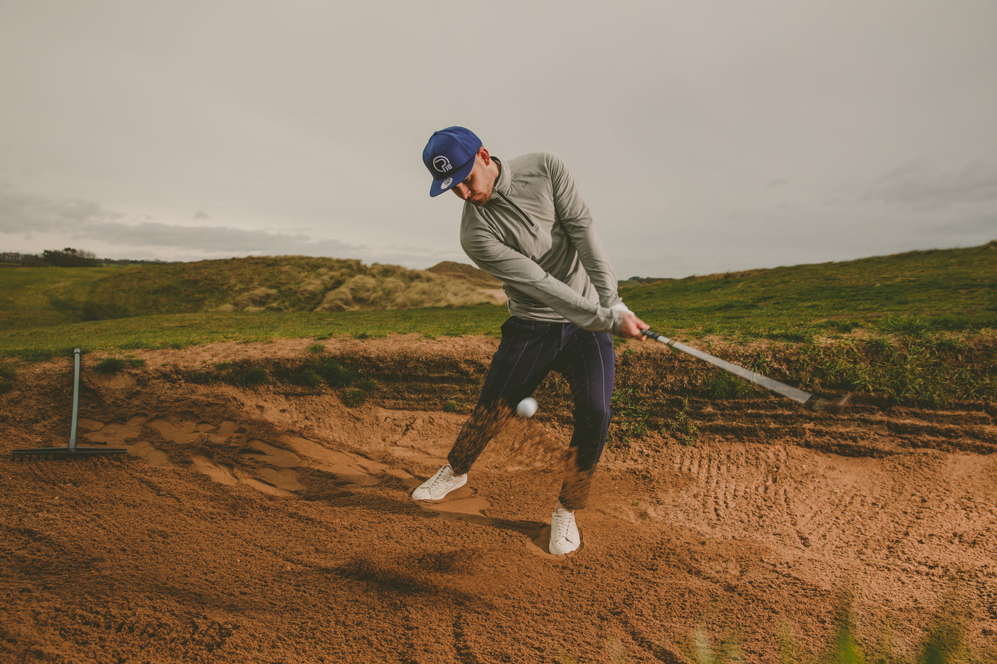 Golfer hitting a bunker shot wearing a blue flat peak Pro18 Sports hat